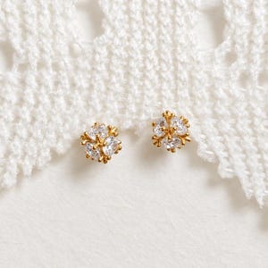May include: A pair of gold stud earrings, each featuring a cluster of small, clear, round-cut stones arranged in a circular, flower-like pattern. The metal settings are polished gold, providing a bright contrast to the sparkling white stones.