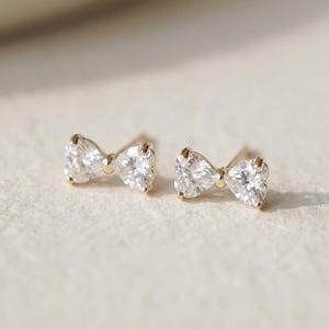 May include: A pair of gold-coloured bow-shaped stud earrings. Each earring features a clear, sparkling stone in the centre of the bow. The earrings are set against a soft, neutral background, highlighting their delicate design.