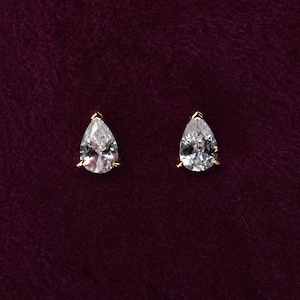 May include: A pair of stud earrings featuring clear, pear-shaped faceted stones set in gold-toned metal prongs. Each earring has a pointed top and a rounded base, showcasing a brilliant cut that reflects light, creating a classic and elegant appearance.