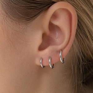 May include: Three silver hoop earrings of varying sizes are worn in the ear. The earrings are small and shiny, reflecting light. The ear is visible against a blurred background.