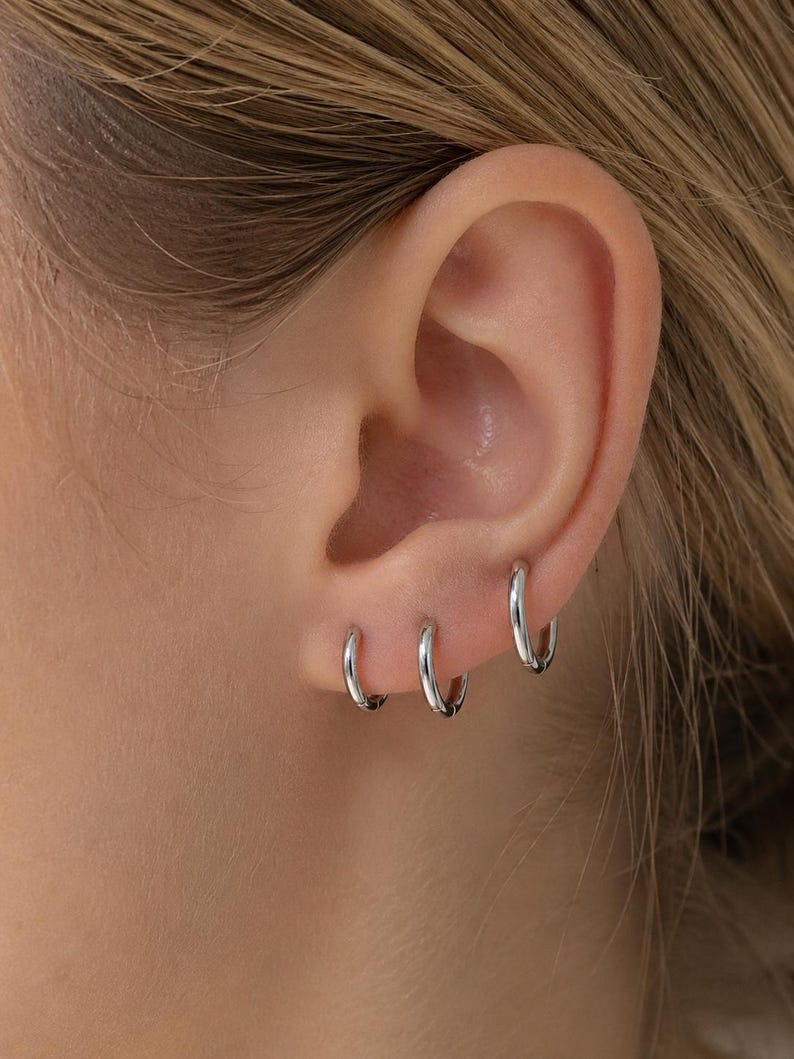May include: Three silver hoop earrings of varying sizes are worn in the ear. The earrings are small and shiny, reflecting light. The ear is visible against a blurred background.
