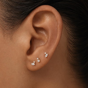 May include: Three gold stud earrings are worn in the earlobe. Each earring features a combination of small, clear, faceted stones in marquise and round cuts, arranged in delicate, asymmetrical clusters that catch the light with a brilliant shine.