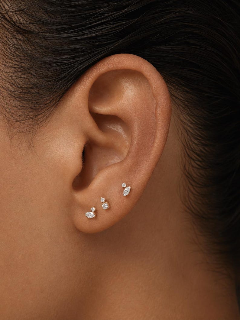 May include: Three gold stud earrings are worn in the earlobe. Each earring features a combination of small, clear, faceted stones in marquise and round cuts, arranged in delicate, asymmetrical clusters that catch the light with a brilliant shine.