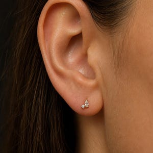 May include: A gold stud earring featuring two clear stones, one small round-cut stone and one larger pear-shaped stone, set in a delicate metal base. The earring is positioned on the lower earlobe, showcasing a minimalist and refined design.