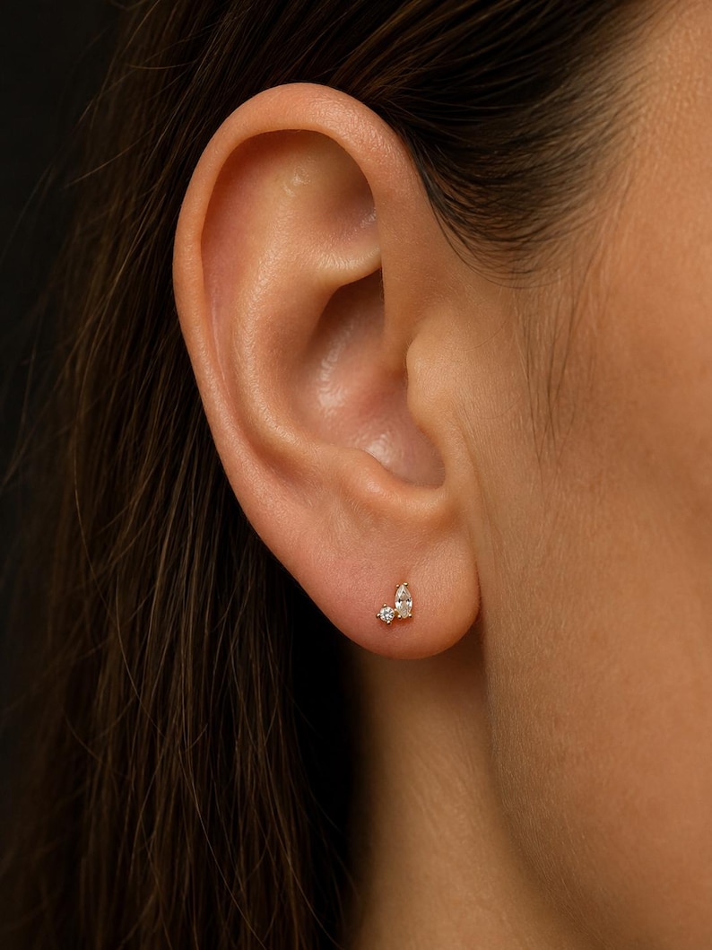 May include: A gold stud earring featuring two clear stones, one small round-cut stone and one larger pear-shaped stone, set in a delicate metal base. The earring is positioned on the lower earlobe, showcasing a minimalist and refined design.