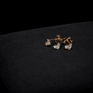 May include: A pair of gold stud earrings, each featuring a marquise-cut clear stone flanked by two smaller round clear stones. The earrings have a post-back closure and are crafted from a polished gold-tone metal, showcasing a cluster design.