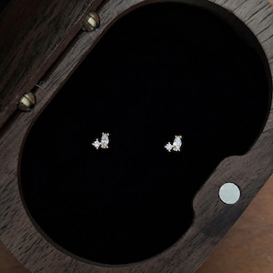 May include: A pair of gold stud earrings, each featuring a small round clear stone paired with a marquise-cut clear stone. The earrings are displayed inside a dark wooden box with a black velvet interior lining, highlighting the metallic gold finish.