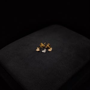 May include: A pair of gold stud earrings, each featuring a cluster of three small, clear, faceted stones arranged in a delicate, asymmetrical pattern. The metal has a polished gold finish, and the stones are set securely in the gold base.