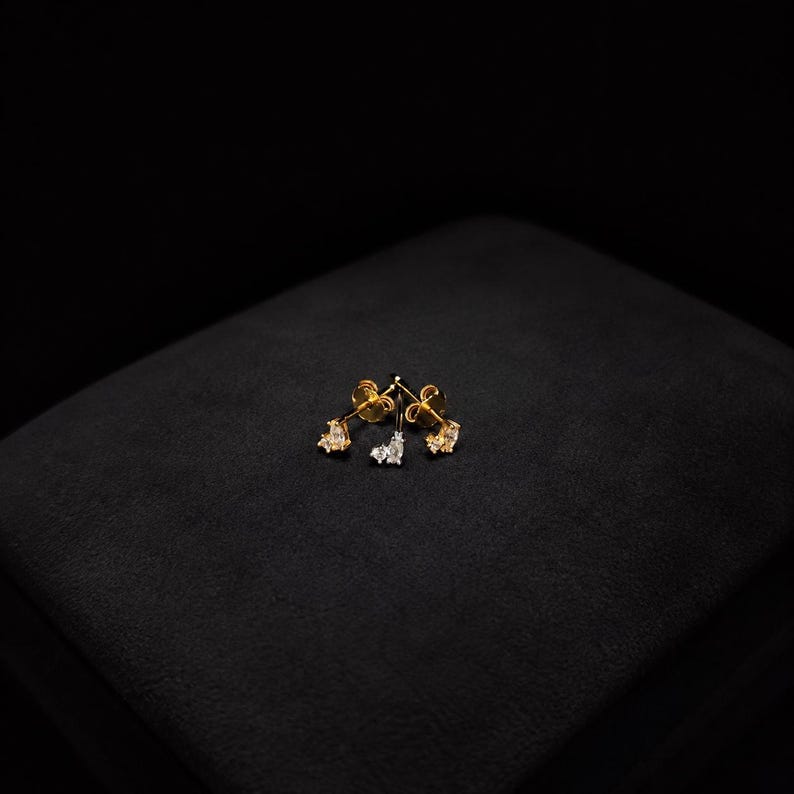 May include: A pair of gold stud earrings, each featuring a cluster of three small, clear, faceted stones arranged in a delicate, asymmetrical pattern. The metal has a polished gold finish, and the stones are set securely in the gold base.