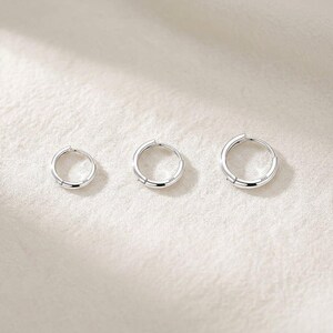 May include: Three silver hoop earrings of varying sizes are arranged on a white, textured surface. The earrings are circular with a simple, polished design. The smallest earring is on the left, with the largest on the right.