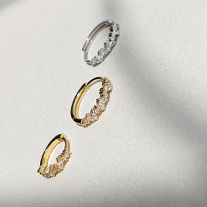 Pode incluir: Três brincos de argola dispostos numa linha diagonal. A argola superior é de metal prateado, enquanto as duas inferiores são de metal dourado. Cada argola apresenta uma fila de pequenas pedras transparentes de corte marquise e redondo.