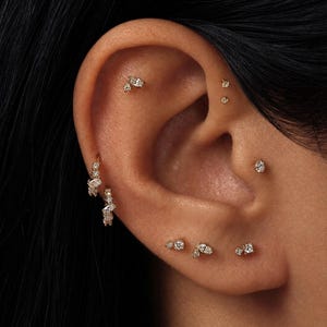 May include: A collection of gold earrings featuring small clear stones, including two hoop earrings on the lobe and several stud earrings placed along the helix, tragus, and earlobe, creating a layered arrangement of delicate jewelry pieces.
