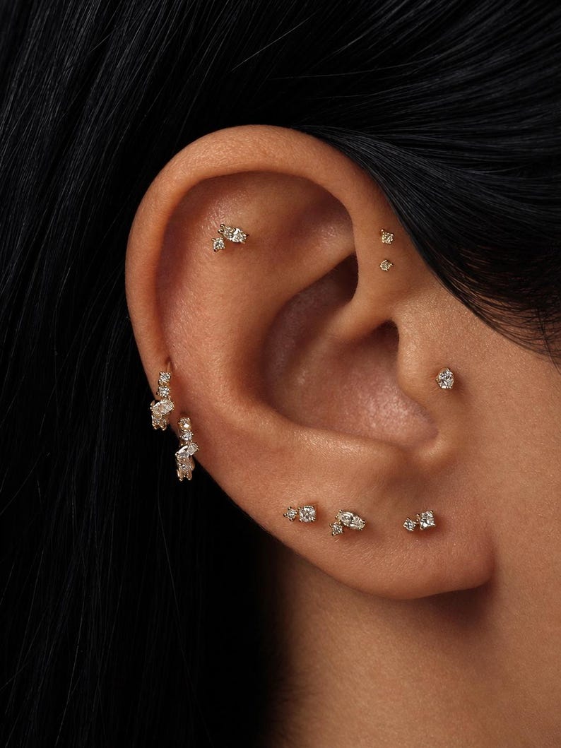 May include: A collection of gold earrings featuring small clear stones, including two hoop earrings on the lobe and several stud earrings placed along the helix, tragus, and earlobe, creating a layered arrangement of delicate jewelry pieces.