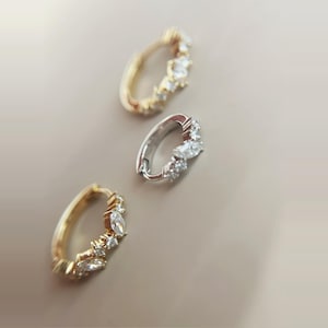 May include: Three small hoop earrings. Two are gold-toned, and one is silver-toned. Each earring is adorned with clear, sparkling gemstones. The earrings are arranged on a neutral-colored surface, showcasing their design.