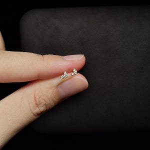 May include: A gold stud earring featuring a cluster of three clear stones, including one marquise-cut stone and two smaller round-cut stones, set in a delicate metal frame. The earring is small and designed for a pierced earlobe.