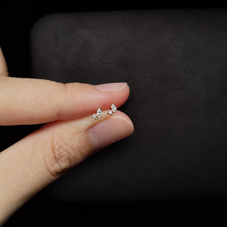 May include: A gold stud earring featuring a cluster of three clear stones, including one marquise-cut stone and two smaller round-cut stones, set in a delicate metal frame. The earring is small and designed for a pierced earlobe.