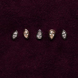 May include: Five individual stud earrings arranged in a horizontal row. The set includes three marquise-cut clear stones in dark metal settings and two pear-cut champagne-coloured stones in gold-toned metal settings, all featuring a bezel design.