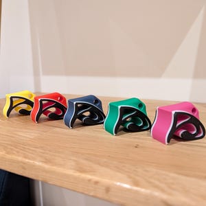 May include: Five colorful, abstract-shaped objects in yellow, red, navy blue, green, and pink, each with a black interior design. They are arranged on a light-colored wooden shelf. The objects have a unique, artistic design.
