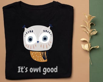 May include: Black t-shirt featuring a cartoon owl design. The owl has large blue eyes, gray and white feathers, and a brown chest. The text "It's owl good" is printed in white below the owl. The shirt is made of a soft material.