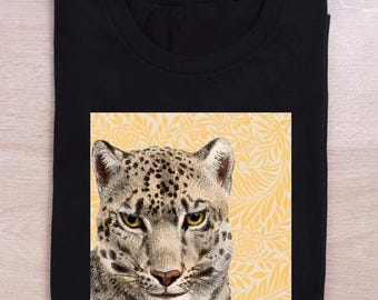 May include: Black t-shirt featuring a detailed illustration of a snow leopard's face. The leopard is set against a yellow floral background. The shirt is folded neatly, showcasing the design prominently.
