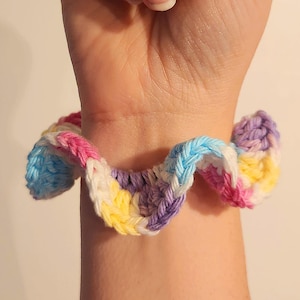 May include: A colorful, crocheted scrunchie in shades of pink, blue, yellow, white, and purple. The handmade hair accessory is wrapped around a wrist, showcasing the vibrant, textured yarn. This scrunchie adds a pop of color to any hairstyle.