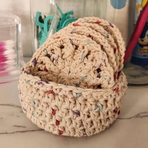 Peut inclure: Deux paniers en crochet beiges, ronds avec un motif texturé et ajouré, empilés l'un sur l'autre. Les paniers ont des mouchetures rouges, bleues et violettes. D'autres articles de salle de bain sont visibles en arrière-plan, sur un comptoir blanc.