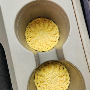 May include: Two round, yellow crocheted coasters sit inside a car's cup holders. The coasters have a textured, floral-like pattern. The cup holders are part of a gray console.