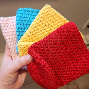 May include: A stack of four handmade crochet washcloths in shades of pink, blue, yellow, and red. The washcloths have a textured appearance and are made with a sparkly yarn. The items are held in a hand.
