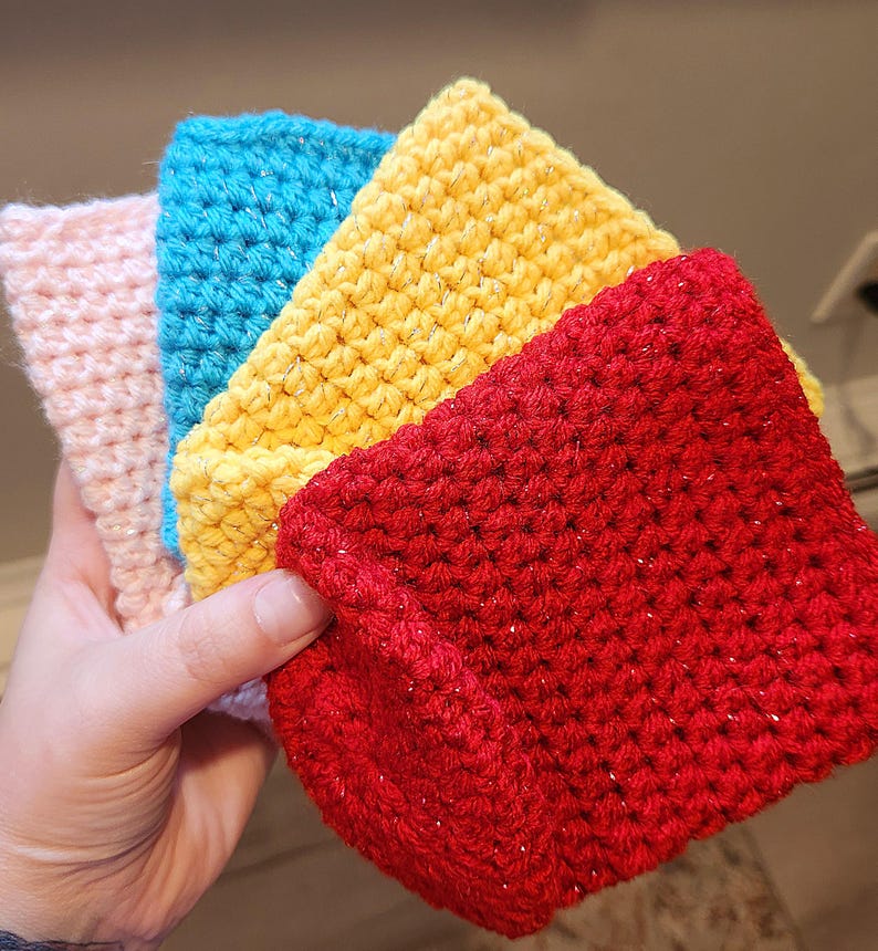 May include: A stack of four handmade crochet washcloths in shades of pink, blue, yellow, and red. The washcloths have a textured appearance and are made with a sparkly yarn. The items are held in a hand.