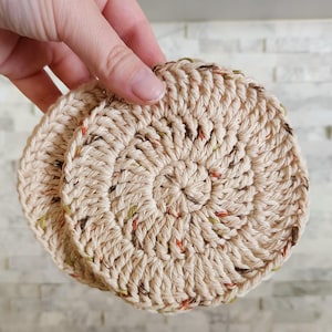 May include: Two beige, circular crochet coasters with flecks of brown, orange, and green. The coasters are held by a hand, showcasing the spiral pattern and textured surface. These handmade coasters are perfect for protecting surfaces.