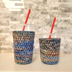 May include: Two clear plastic cups with red straws, each wrapped in a colorful, crocheted cozy. The cozies feature a mix of blue, orange, gray, and white yarn. The cups are on a white surface against a textured, light-colored wall.