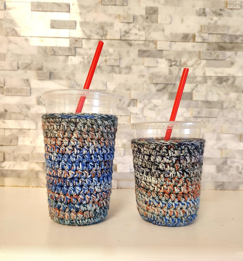May include: Two clear plastic cups with red straws, each wrapped in a colorful, crocheted cozy. The cozies feature a mix of blue, orange, gray, and white yarn. The cups are on a white surface against a textured, light-colored wall.