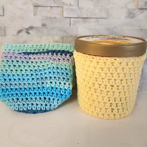 May include: Two crocheted cozies. One is a light yellow color, and the other is a mix of blue, green, and purple. The yellow cozy is around a container. The other cozy is empty and has a rounded shape.