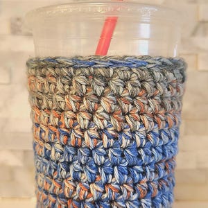 May include: A clear plastic cup with a red straw, encased in a handmade crochet cozy. The cozy features a textured pattern in shades of blue, orange, and gray, providing insulation and a stylish look.