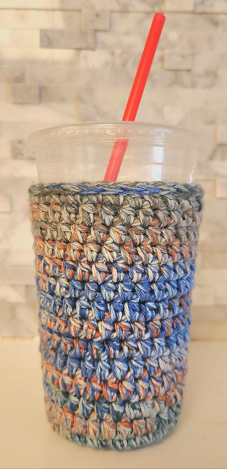May include: A clear plastic cup with a red straw, encased in a handmade crochet cozy. The cozy features a textured pattern in shades of blue, orange, and gray, providing insulation and a stylish look.