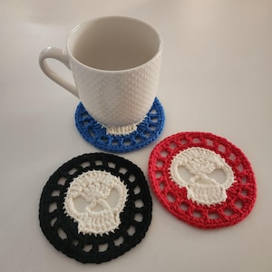 May include: A white ceramic mug rests on a blue crocheted coaster. Two additional crocheted coasters are visible: one black and one red, each featuring a white skull design. The coasters are handmade and have an open, lacy pattern.