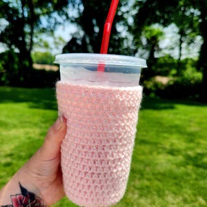 May include: A clear plastic cup with a lid and a red straw, held in a pink crocheted cozy. The cozy is wrapped around the cup, providing insulation. The background is a blurred view of green trees and grass.