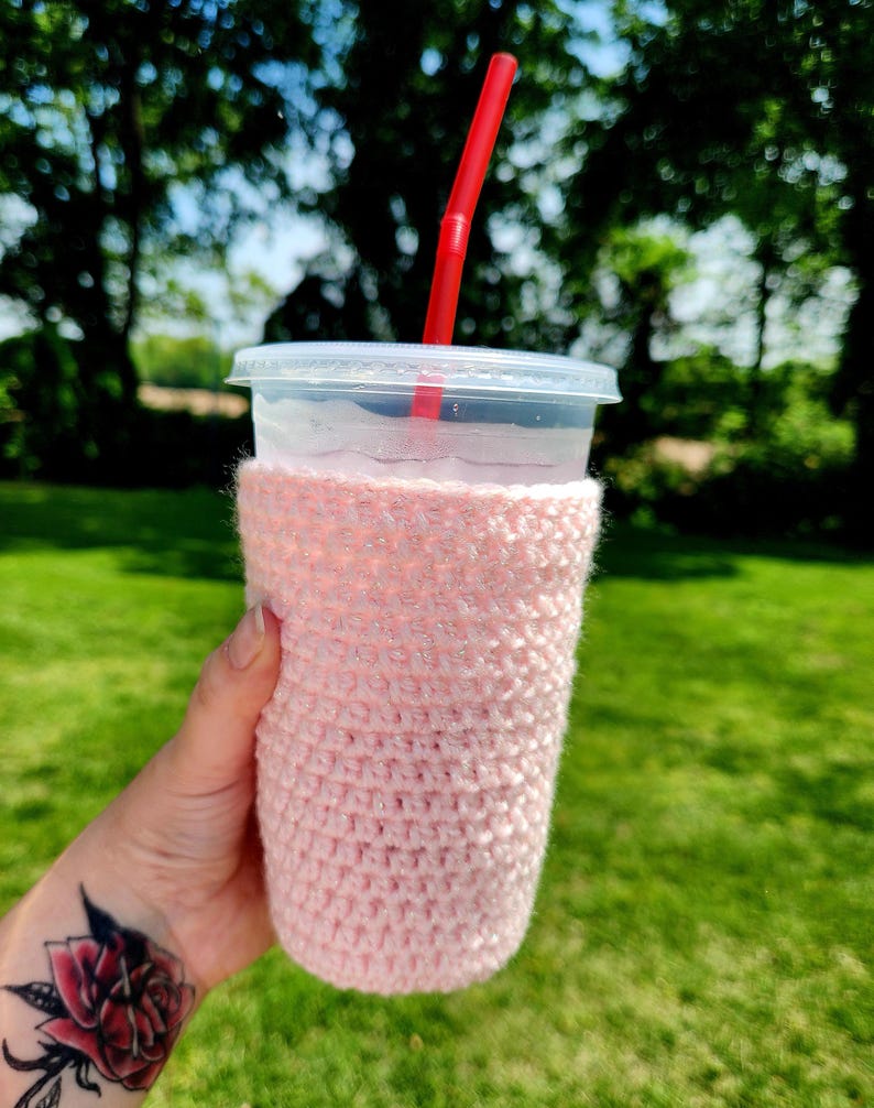 May include: A clear plastic cup with a lid and a red straw, held in a pink crocheted cozy. The cozy is wrapped around the cup, providing insulation. The background is a blurred view of green trees and grass.