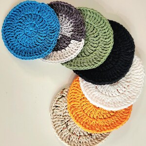 May include: A collection of handmade crochet coasters in various colors, including blue, green, black, white, orange, and beige. The coasters are round and feature a textured, spiral design. These coasters are perfect for protecting surfaces from drink rings.