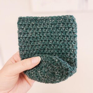 May include: A hand-crocheted, dark green cozy with flecks of white yarn. The cozy is rectangular with a rounded bottom, and is designed to hold a small object. The yarn has a textured appearance.