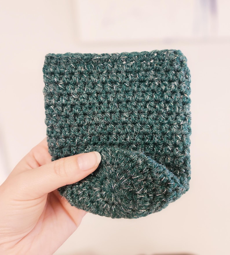 May include: A hand-crocheted, dark green cozy with flecks of white yarn. The cozy is rectangular with a rounded bottom, and is designed to hold a small object. The yarn has a textured appearance.