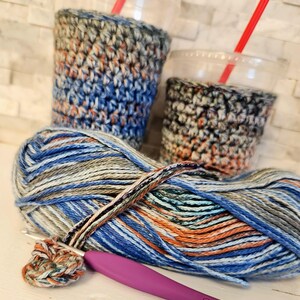 May include: Two clear plastic cups with red straws are encased in handmade crochet cozies. A ball of multicolored yarn with blue, orange, and gray hues sits next to a crochet hook and a small crocheted circle.