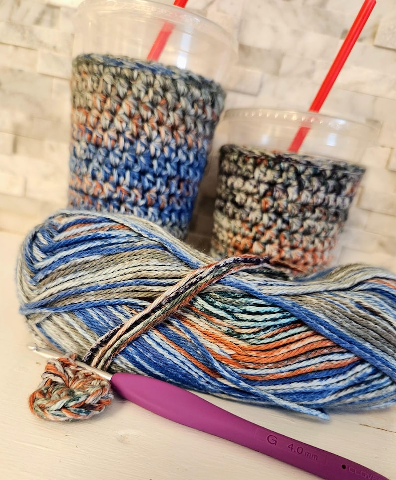 May include: Two clear plastic cups with red straws are encased in handmade crochet cozies. A ball of multicolored yarn with blue, orange, and gray hues sits next to a crochet hook and a small crocheted circle.