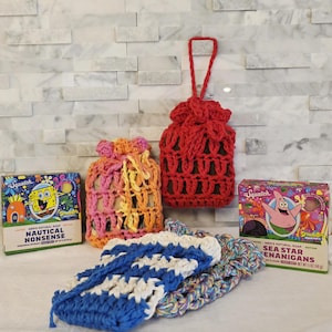 May include: An assortment of handmade crochet items, including soap savers in red, orange, and blue, and soap pouches. Two boxes of "Squatch" brand soap are also visible, featuring cartoon characters and the phrases "Nautical Nonsense" and "Sea Star Enanigans."