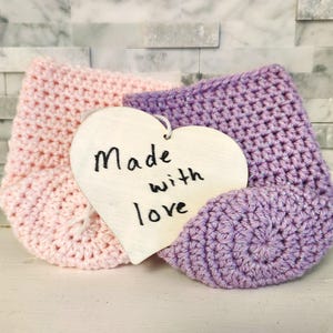 May include: A pair of handmade crochet baby booties in light pink and lavender, with a wooden heart-shaped tag that reads "Made with love". The booties are displayed on a white surface against a textured background.