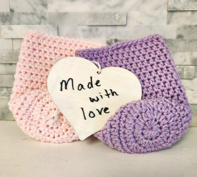May include: A pair of handmade crochet baby booties in light pink and lavender, with a wooden heart-shaped tag that reads "Made with love". The booties are displayed on a white surface against a textured background.