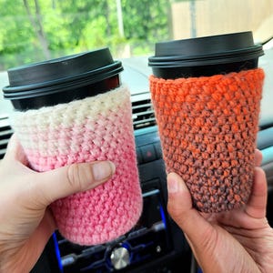 May include: Two coffee cups with black lids are held in the image. Each cup is wrapped in a crocheted cozy. One cozy is pink and white, the other is orange and brown. The cozies are designed to insulate the cups.