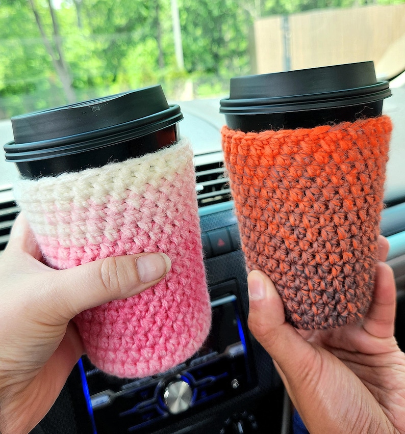 May include: Two coffee cups with black lids are held in the image. Each cup is wrapped in a crocheted cozy. One cozy is pink and white, the other is orange and brown. The cozies are designed to insulate the cups.