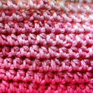 May include: Close-up of a handmade crochet fabric with a gradient of pink and red hues. The yarn has a subtle metallic silver thread woven throughout, adding a touch of sparkle. The texture is dense and shows the individual stitches.
