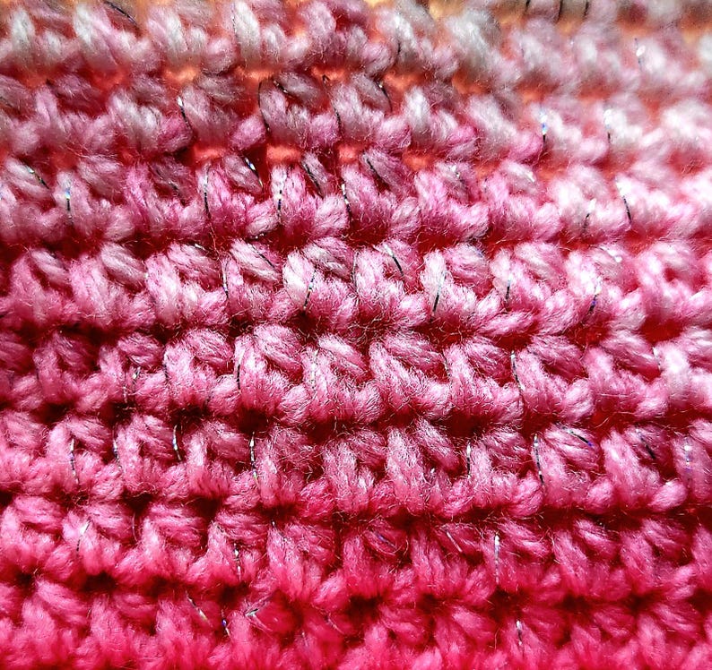 May include: Close-up of a handmade crochet fabric with a gradient of pink and red hues. The yarn has a subtle metallic silver thread woven throughout, adding a touch of sparkle. The texture is dense and shows the individual stitches.